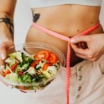 Woman holding vegetable salad and measuring waist with tape, symbolizing healthy lifestyle choices.
